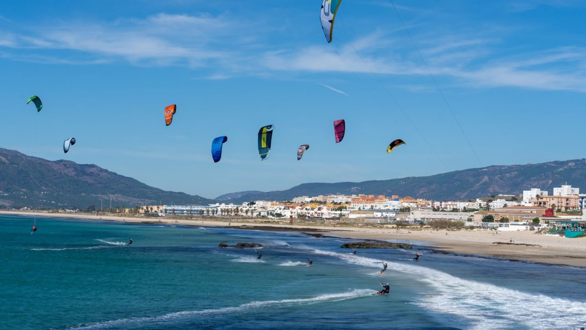 Colorful kites soar above a sandy beach with riders engaging in kitesurfing along the coastline.
