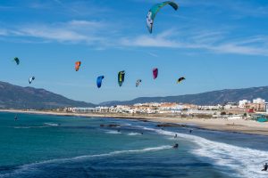 Colorful kites soar above a sandy beach with riders engaging in kitesurfing along the coastline.