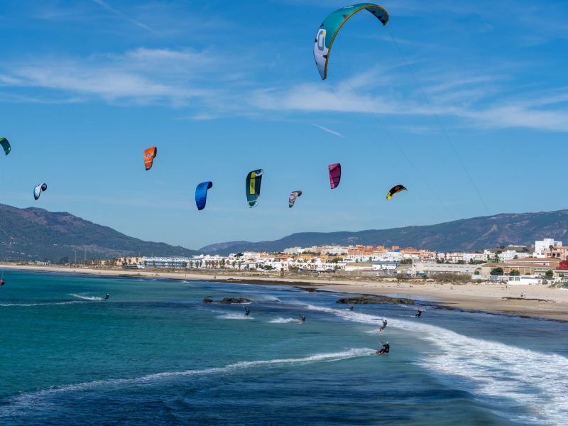 Colorful kites soar above a sandy beach with riders engaging in kitesurfing along the coastline.