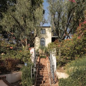 Casa Ivarene, a villa in Hollywood Hills, features lush gardens, a brick pathway, and vibrant flowers surrounding the entrance.