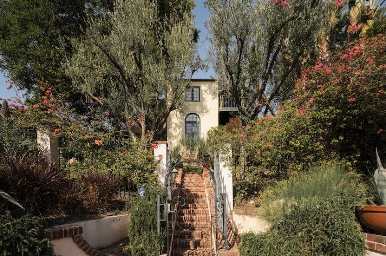 Casa Ivarene, a villa in Hollywood Hills, features lush gardens, a brick pathway, and vibrant flowers surrounding the entrance.