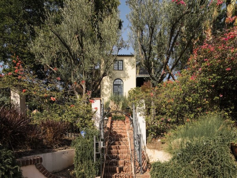 Casa Ivarene, a villa in Hollywood Hills, features lush gardens, a brick pathway, and vibrant flowers surrounding the entrance.