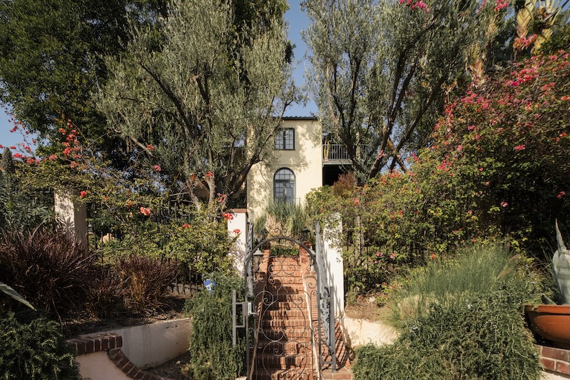 Casa Ivarene, a villa in Hollywood Hills, features lush gardens, a brick pathway, and vibrant flowers surrounding the entrance.