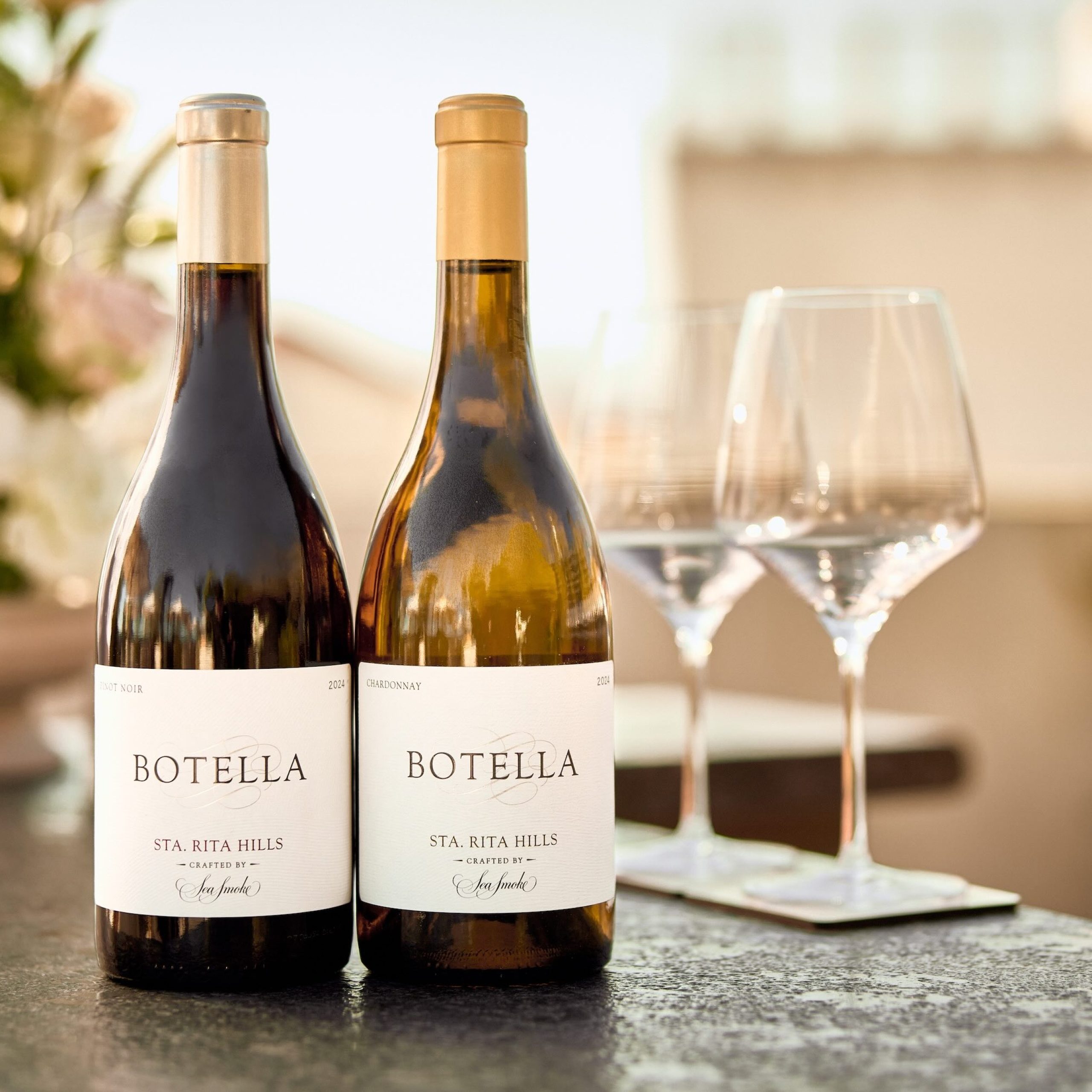 Two bottles of Sea Smoke Botella wine with glasses in the background, set on a dark stone surface.