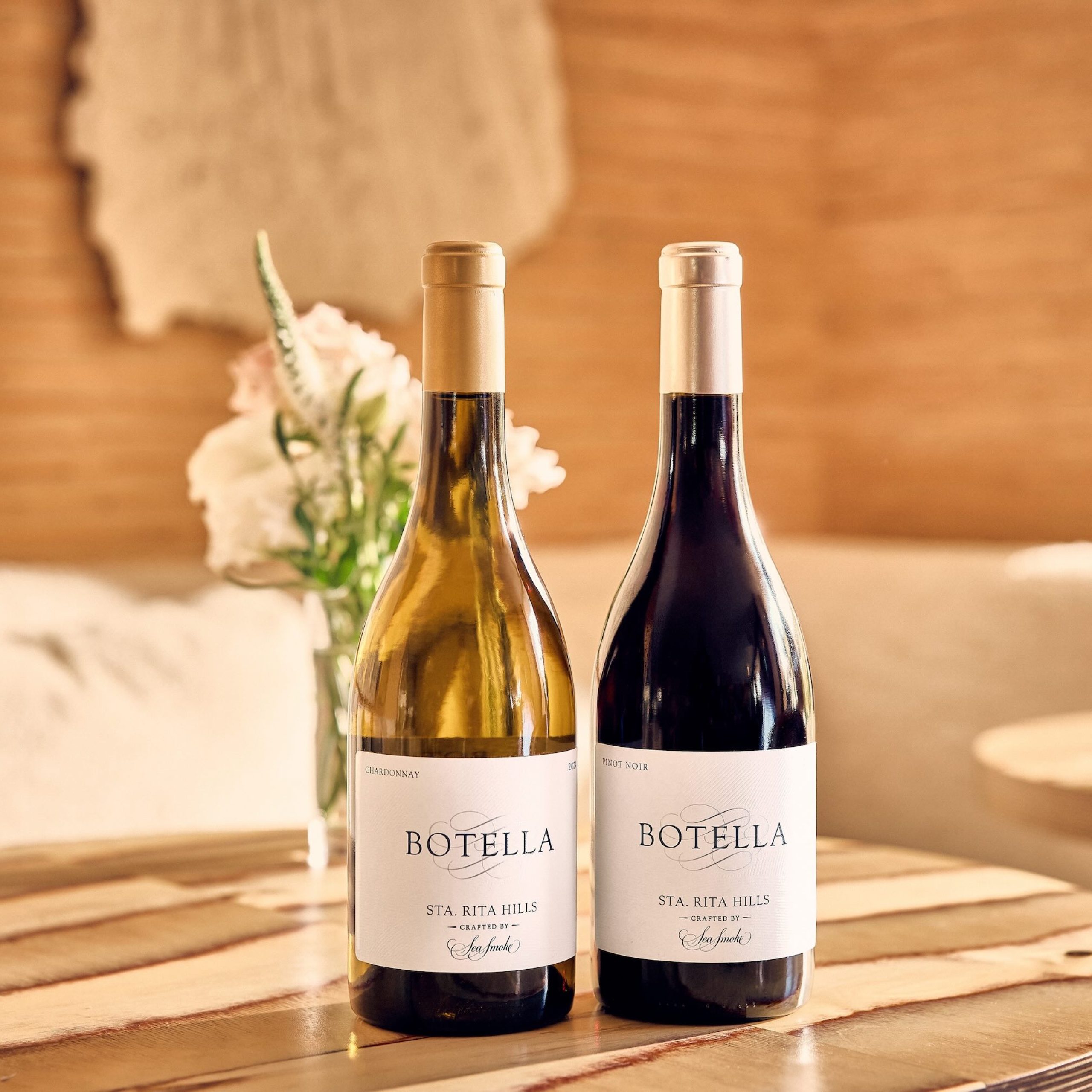 Two bottles of Sea Smoke's Botella wine, one Chardonnay and one Pinot Noir, displayed on a wooden table with flowers in the background.