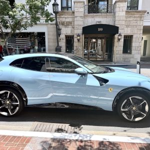 The Ferrari Purosangue in a light blue color showcases its sleek design and luxury features on a sunlit street.