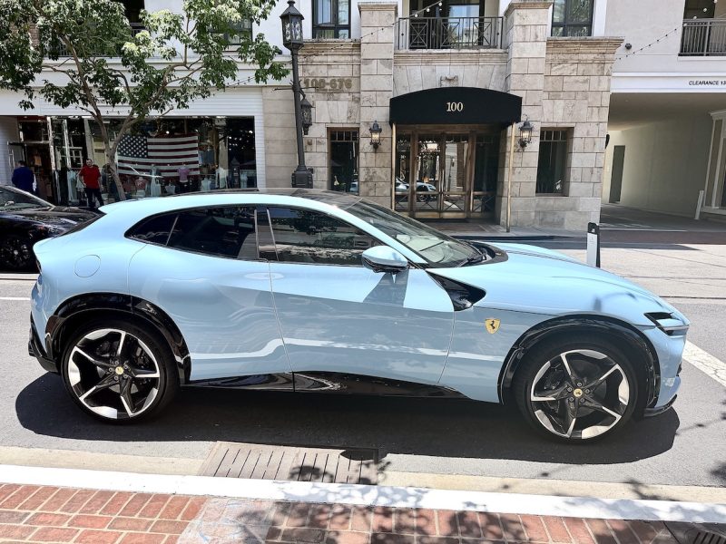 The Ferrari Purosangue in a light blue color showcases its sleek design and luxury features on a sunlit street.