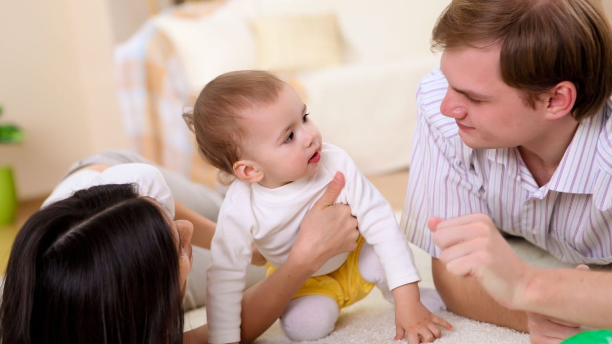 A baby plays on a soft rug with a smiling parent on each side, emphasizing joyful family interactions in a cozy home setting.