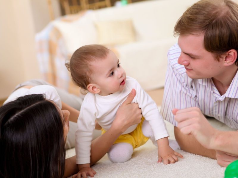 A baby plays on a soft rug with a smiling parent on each side, emphasizing joyful family interactions in a cozy home setting.