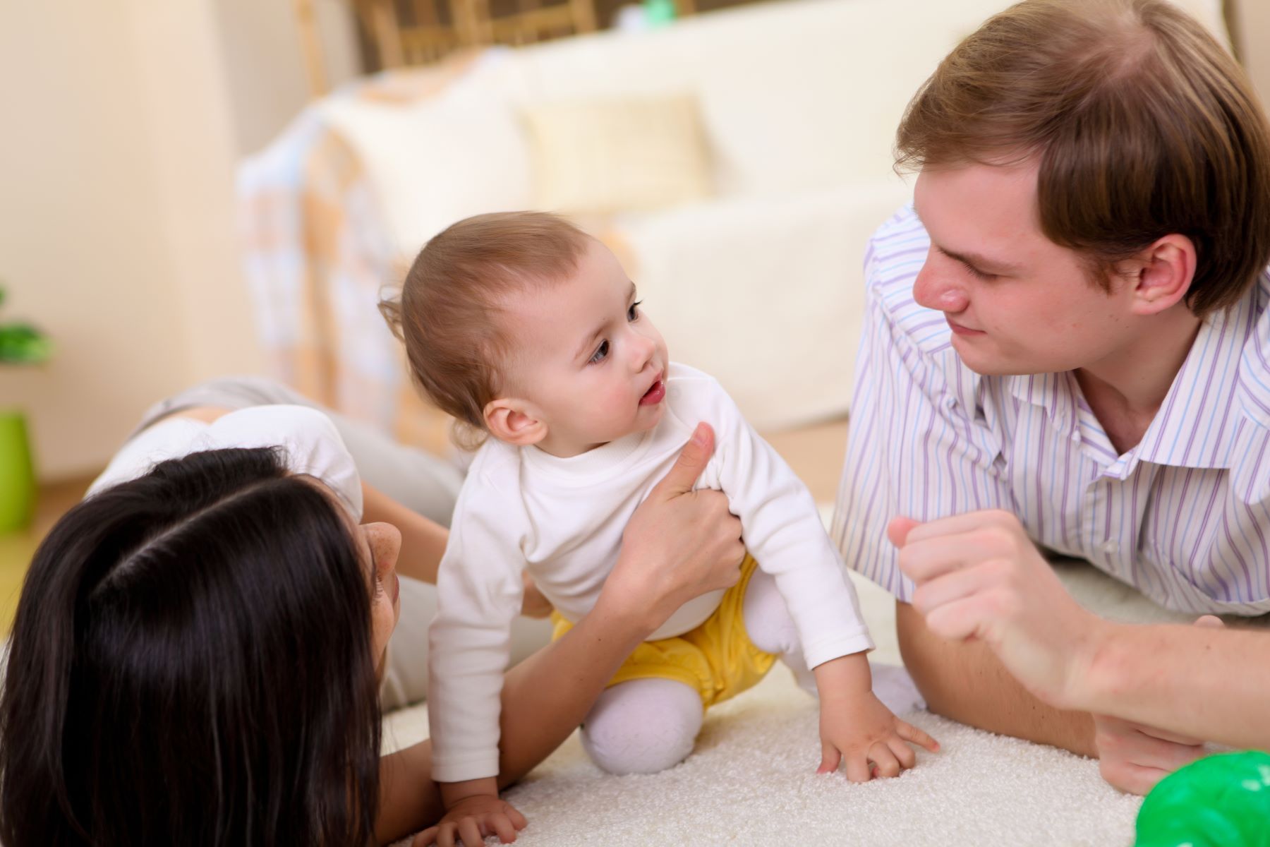 A baby plays on a soft rug with a smiling parent on each side, emphasizing joyful family interactions in a cozy home setting.