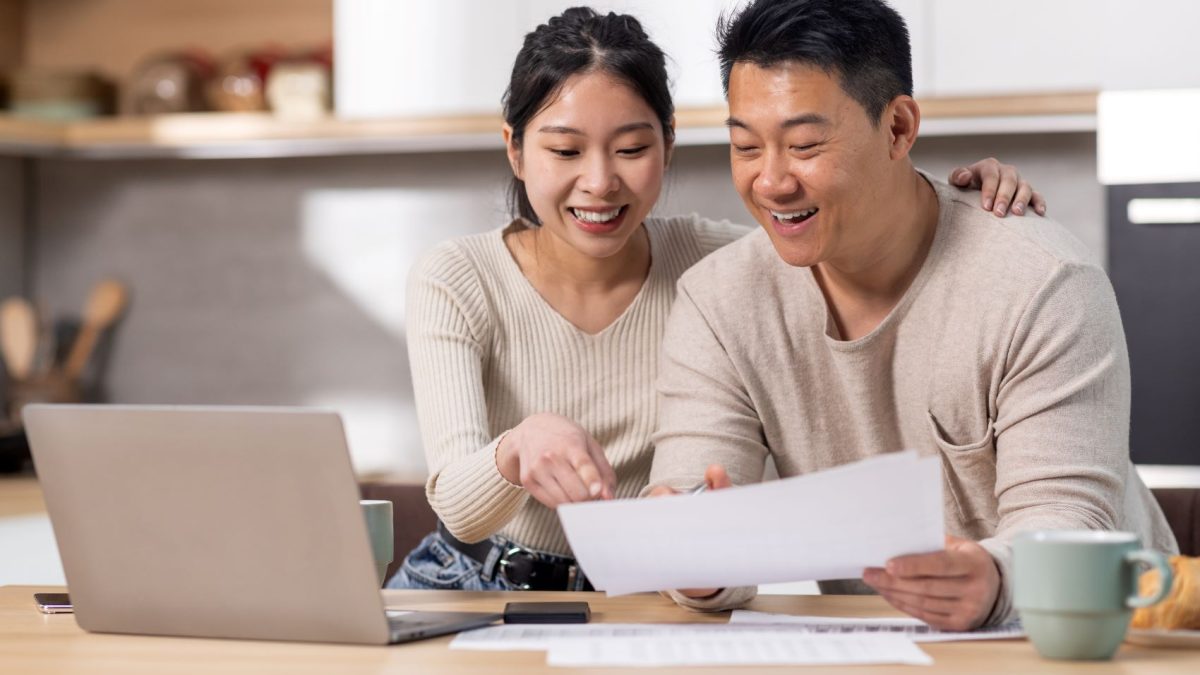 A couple reviews financial documents together at a desk, smiling and engaged, with a laptop and coffee nearby.
