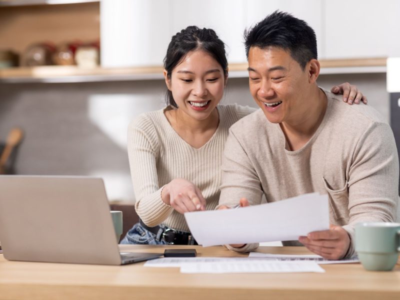 A couple reviews financial documents together at a desk, smiling and engaged, with a laptop and coffee nearby.