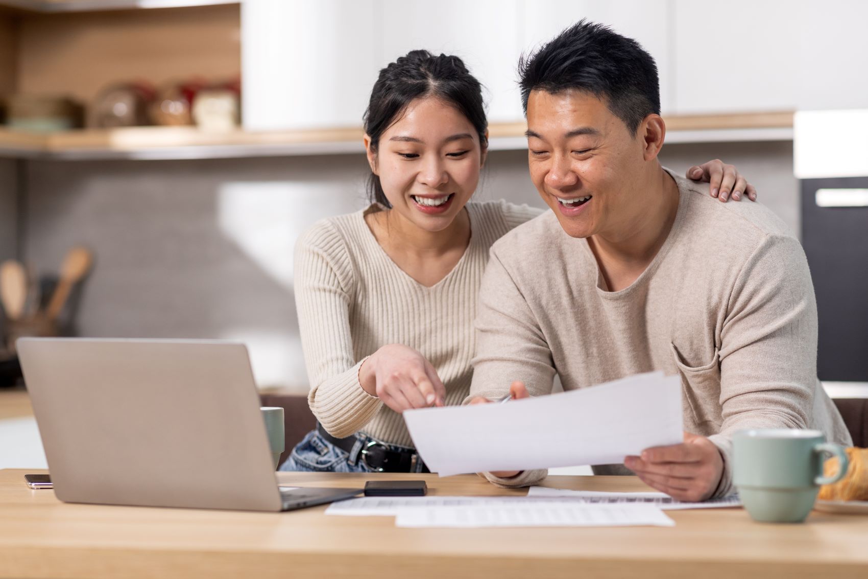 A couple reviews financial documents together at a desk, smiling and engaged, with a laptop and coffee nearby.