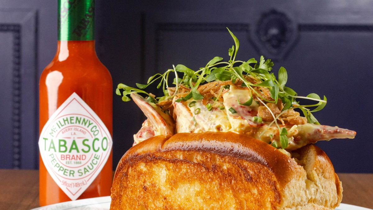 Lobster roll topped with greens and served with a bottle of TABASCO sauce on a plate at a restaurant.