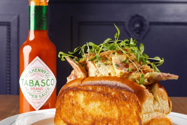 Lobster roll topped with greens and served with a bottle of TABASCO sauce on a plate at a restaurant.