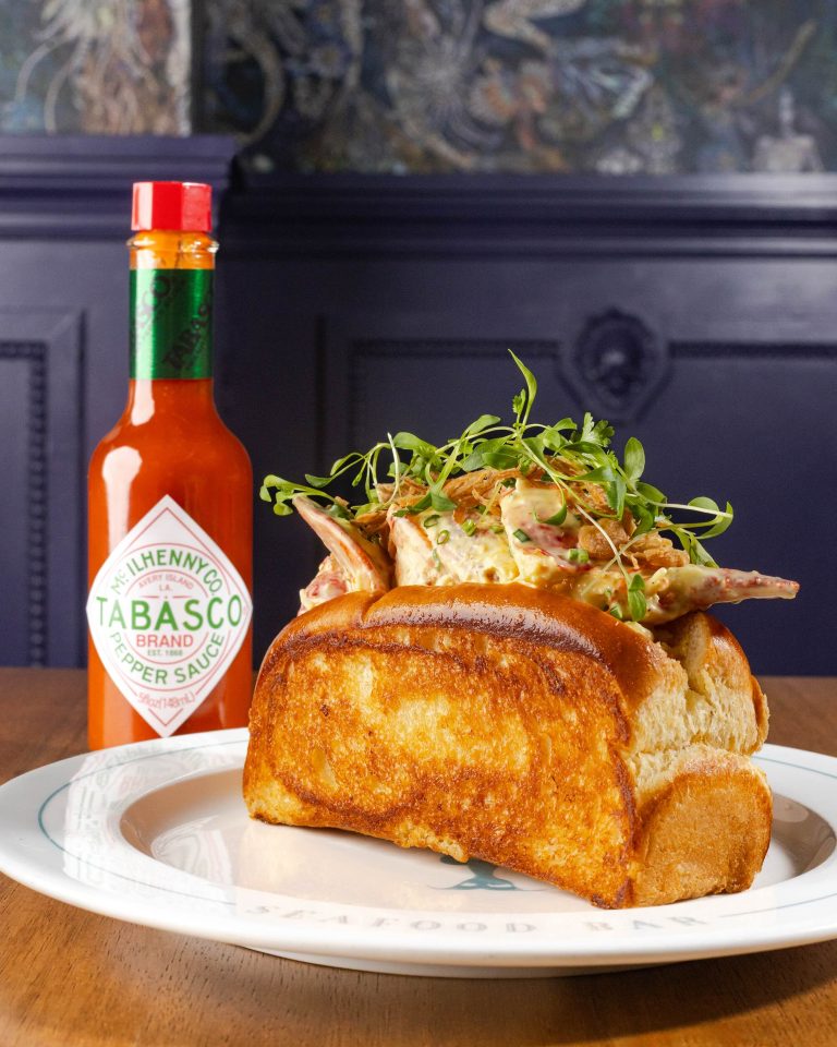 Lobster roll topped with greens and served with a bottle of TABASCO sauce on a plate at a restaurant.