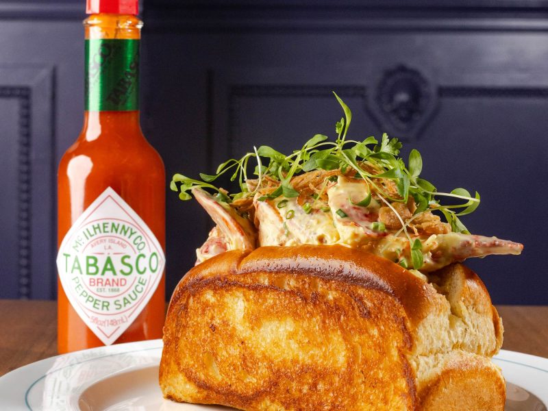 Lobster roll topped with greens and served with a bottle of TABASCO sauce on a plate at a restaurant.
