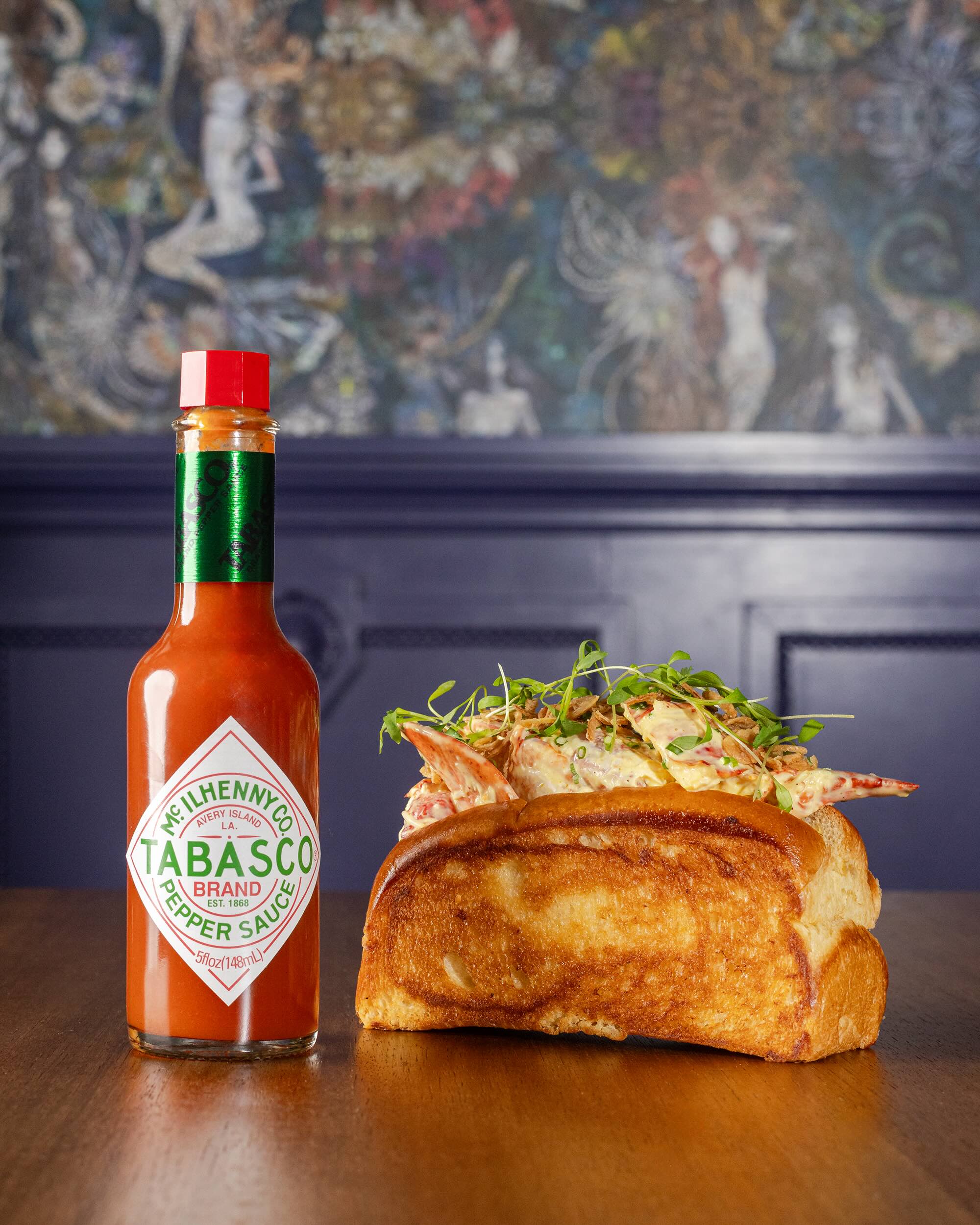 A lobster roll topped with greens, paired with a bottle of TABASCO sauce, set against a vibrant background.