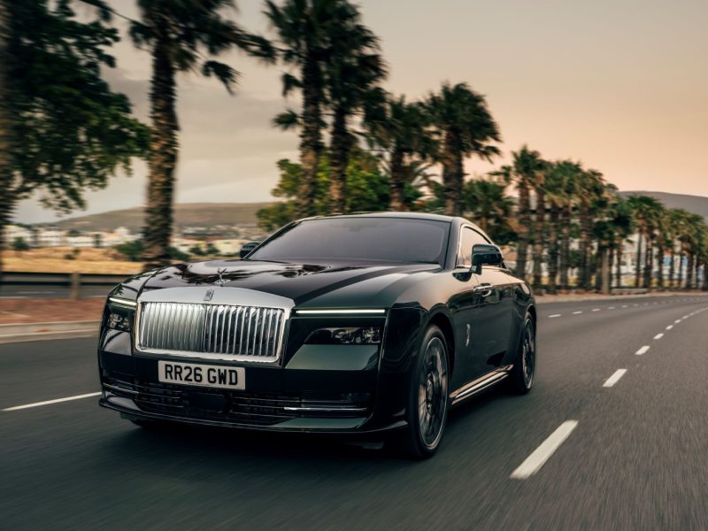 Rolls-Royce Spectre Series II driving on a palm-lined road at sunset, showcasing its luxury design and powerful presence.