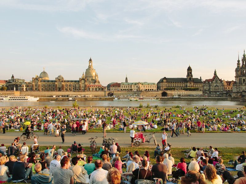 Crowds enjoying an outdoor event along the Elbe River with Dresden's historic skyline in the background.