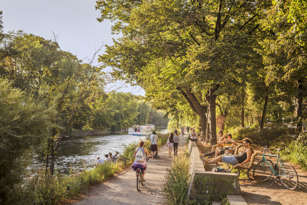 People enjoy a sunny day along the Landwehr Canal in Berlin, with cyclists, walkers, and benches by the water.