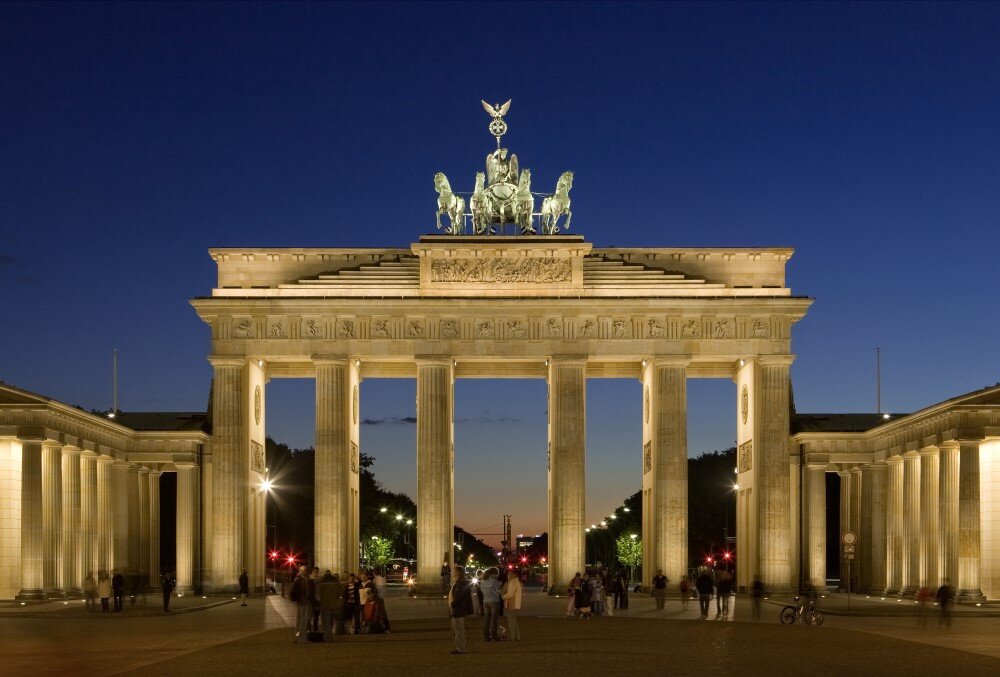 Brandenburg Gate illuminated at dusk, showcasing its historic architecture and vibrant atmosphere with people strolling nearby.