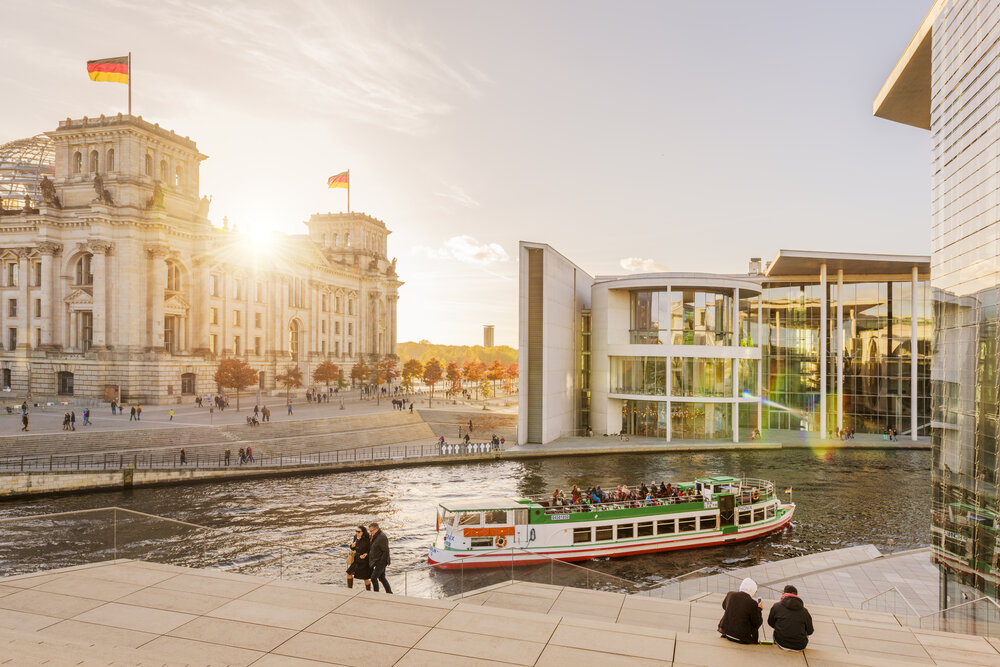 The sun sets over Berlin's government district, with people along the Spree River and a boat passing by.