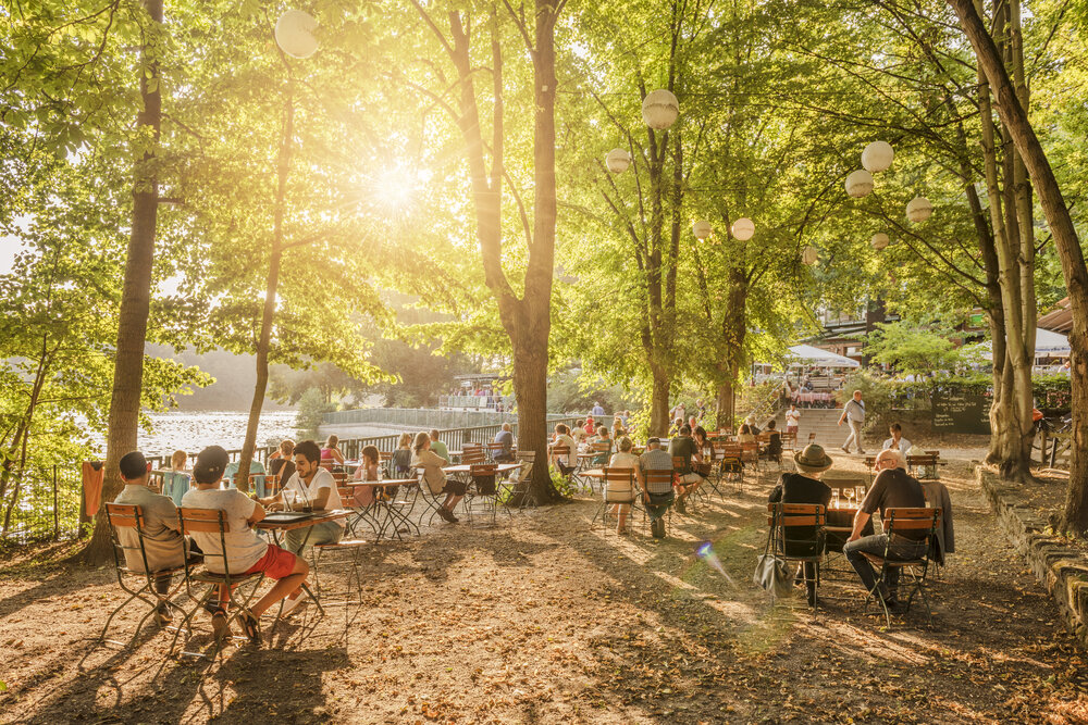 Sunlit outdoor café by a lake, with people dining at tables among green trees and hanging lanterns in Berlin.