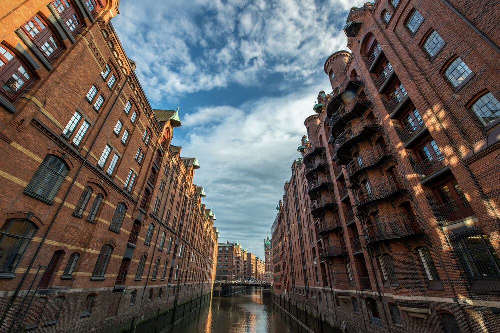 Historic red-brick warehouses line a canal in Speicherstadt, Hamburg, under a partly cloudy sky.