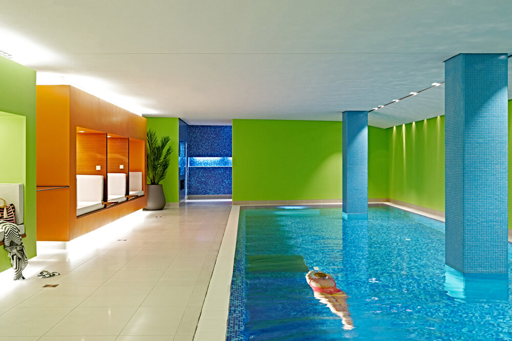 Indoor spa pool with vibrant green walls and wooden rest areas, featuring a person swimming in crystal-clear water.