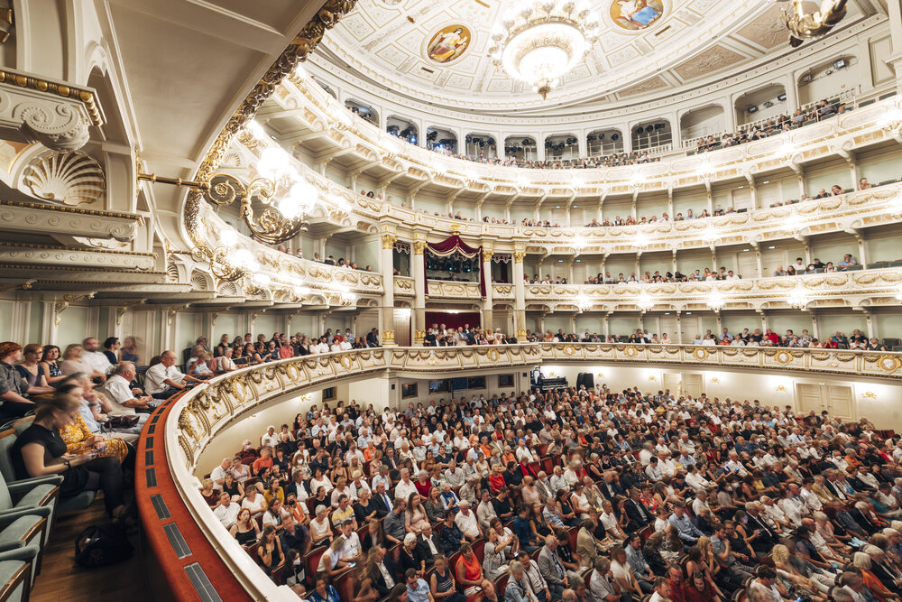 View of a packed audience at the Semperoper in Dresden during an evening performance, showcasing its ornate interior and architecture.