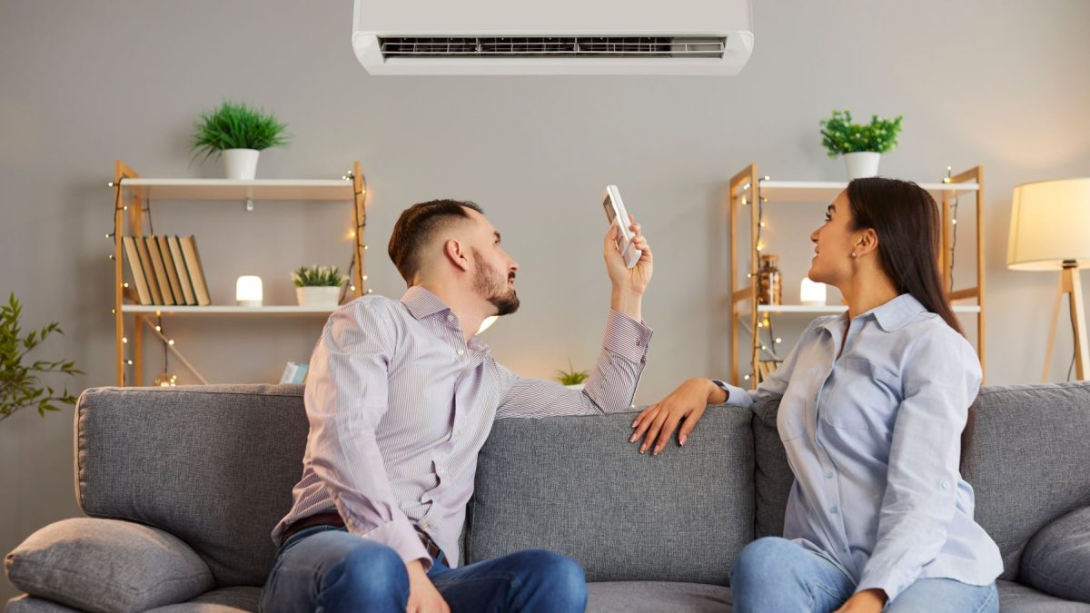 A couple sits on a gray couch, with one person using a remote to adjust a high-end mini-split air conditioner above them.