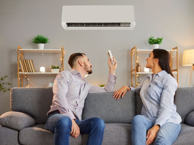 A couple sits on a gray couch, with one person using a remote to adjust a high-end mini-split air conditioner above them.