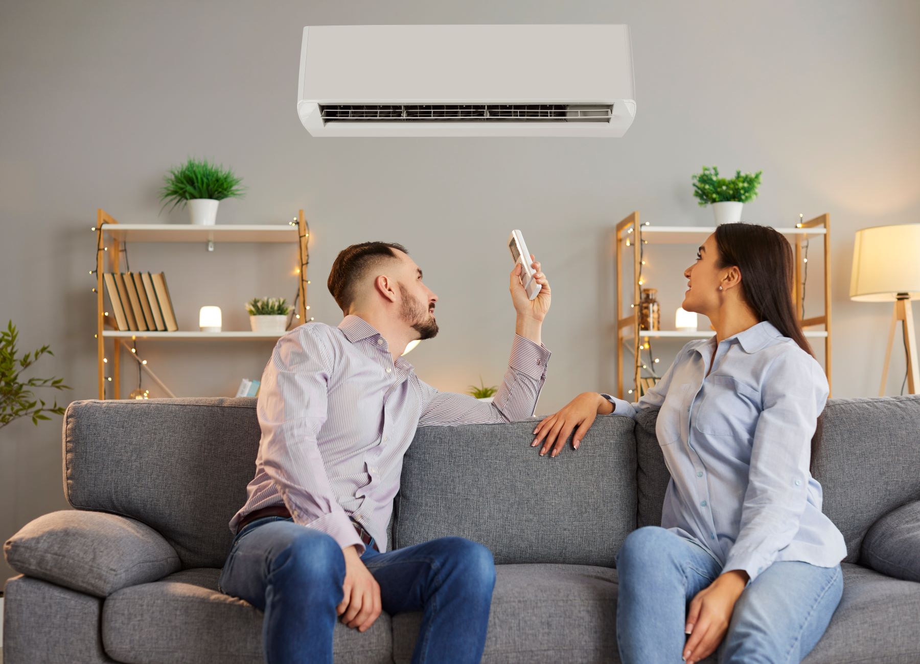 A couple sits on a gray couch, with one person using a remote to adjust a high-end mini-split air conditioner above them.