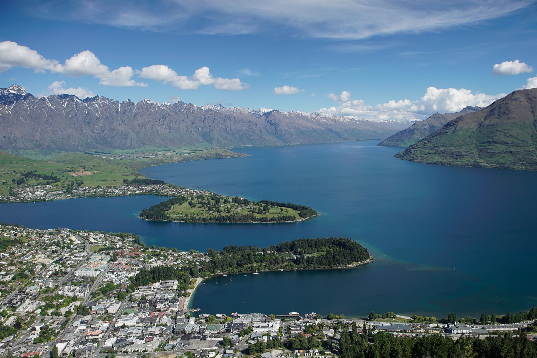A sweeping view of Queenstown, New Zealand, featuring lakes, islands, and mountains under a clear blue sky.