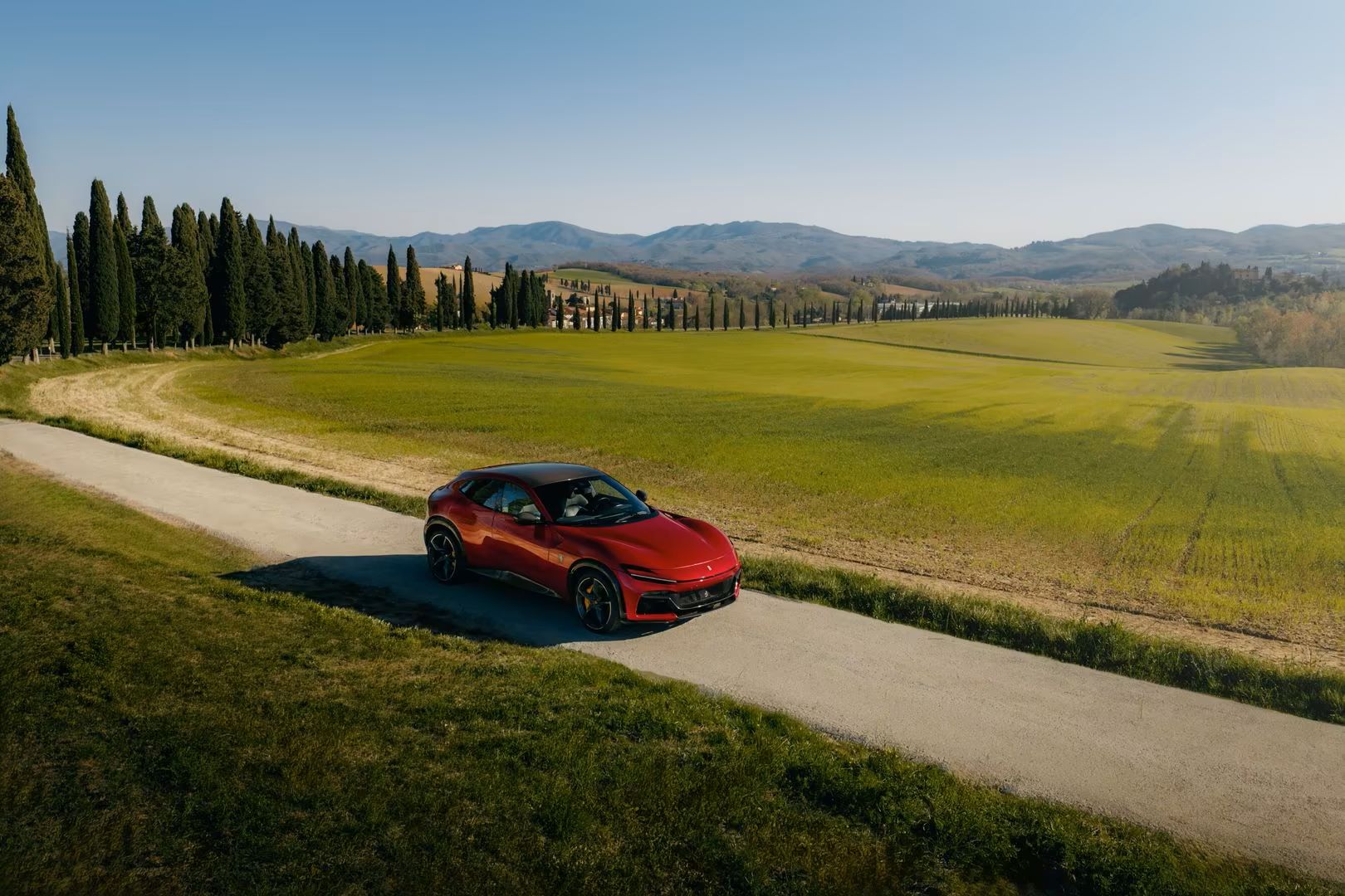 Ferrari Purosangue parked on a country road, surrounded by green fields and tall trees under a clear blue sky.