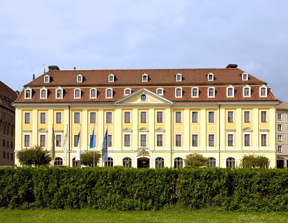 Elegant yellow building with a distinctive roof and several flags outside, surrounded by green landscaping.