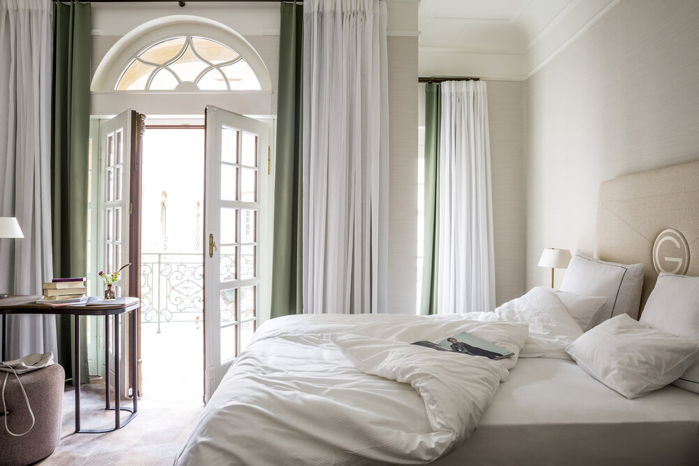 Bright and airy seaside hotel room featuring a bed, round table with books, and open French doors leading to a balcony.