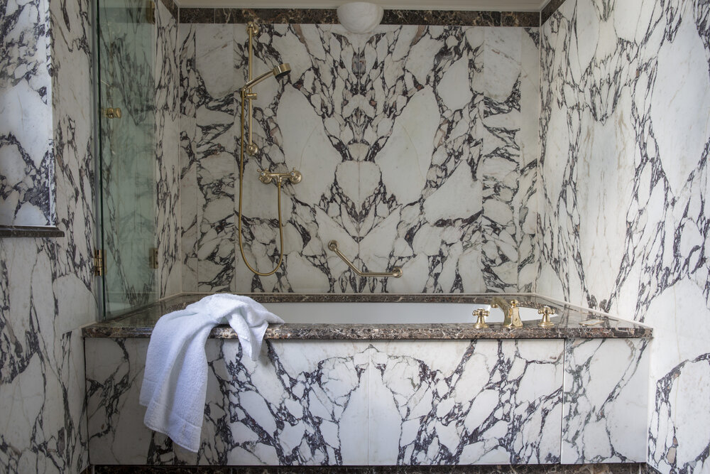 Luxurious bathroom featuring marble walls, a soaking tub, and elegant gold fixtures. A white towel is draped over the tub edge.