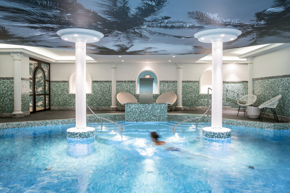 Indoor swimming pool area with turquoise tiles, palm tree ceiling design, and a person swimming in the water.
