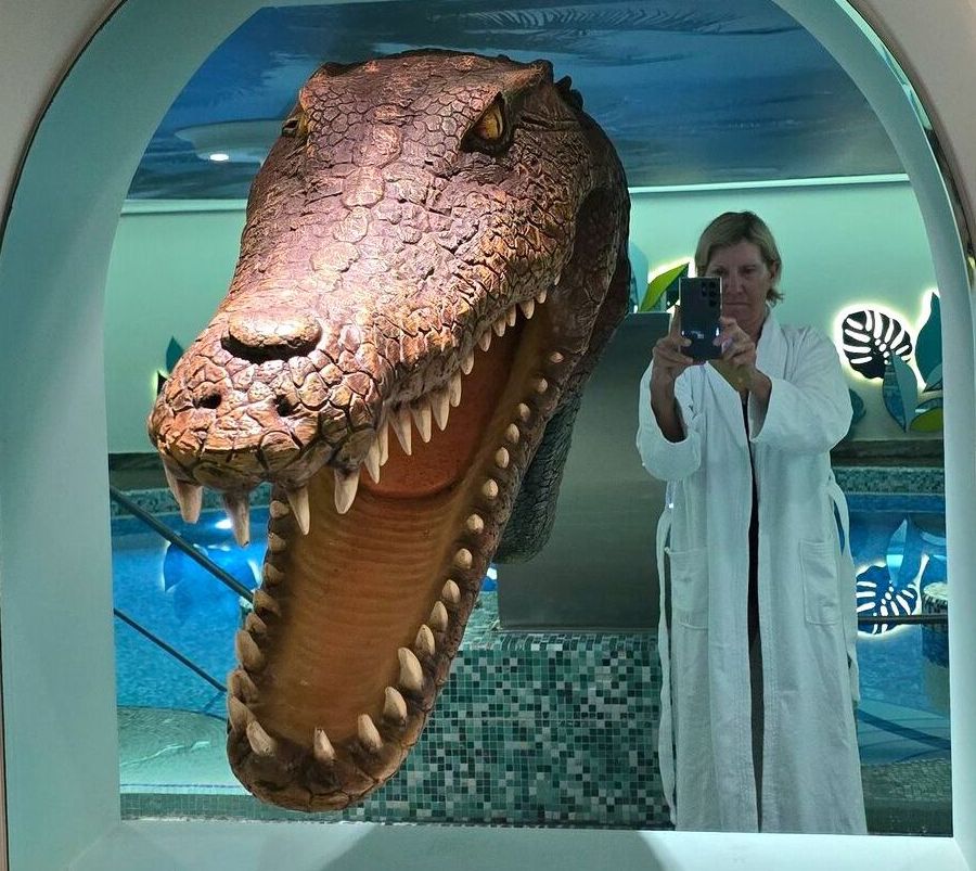 Woman in a bathrobe taking a selfie in front of a large crocodile sculpture, with a blue pool in the background.