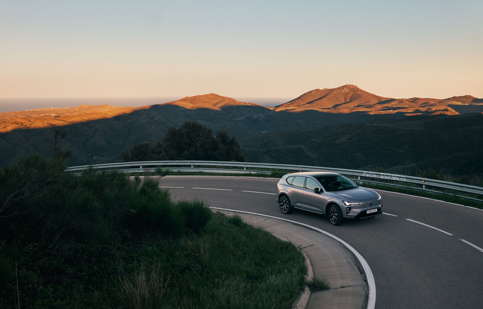 The Volvo EX60 navigates a curving mountain road, framed by rugged hills and a soft sunset glow.