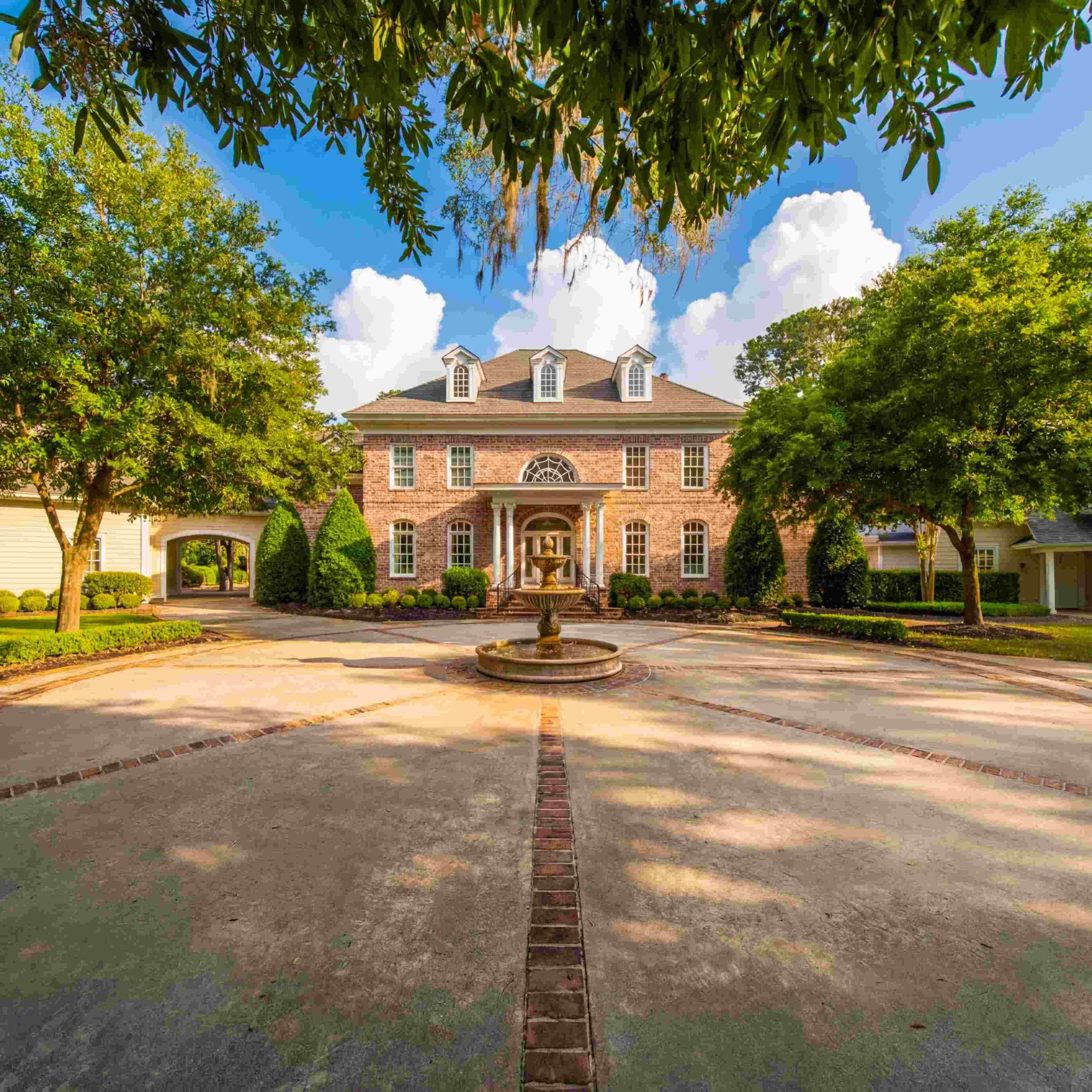 Elegant estate in Bluffton featuring a circular driveway, fountain, and lush landscaping under a bright blue sky.