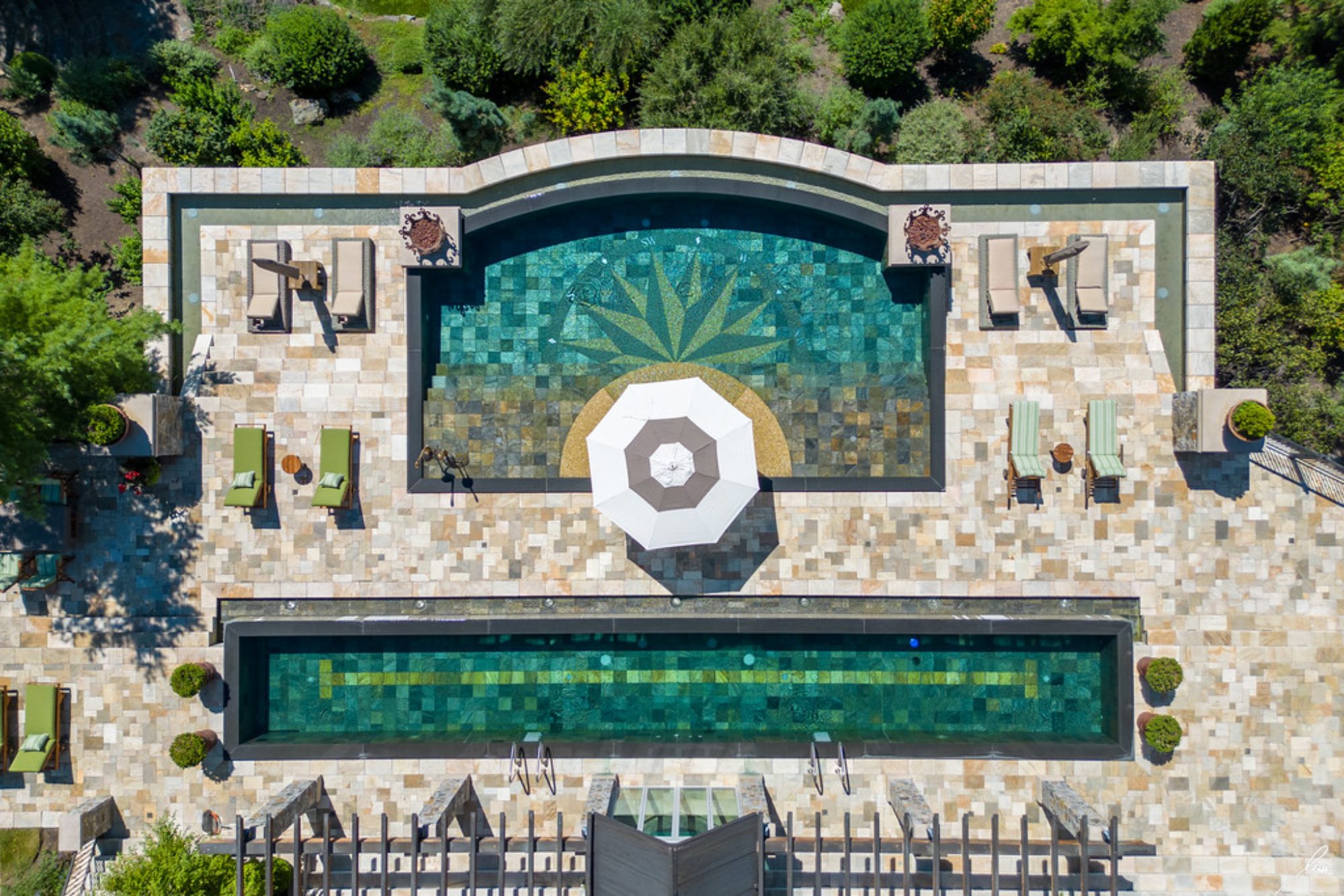 Aerial view of luxurious pools and lounging area surrounded by lush greenery at an exclusive waterfront estate.