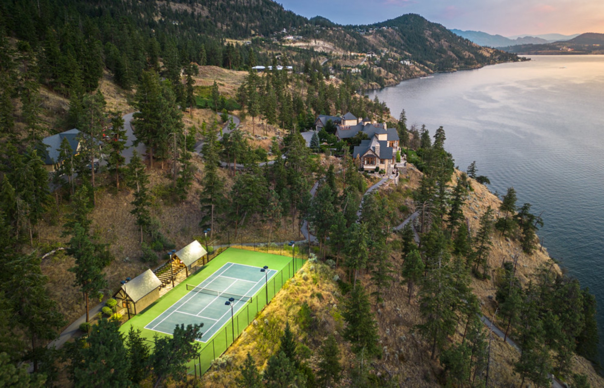 Aerial view of a private tennis court near a luxury waterfront estate surrounded by trees and hills.