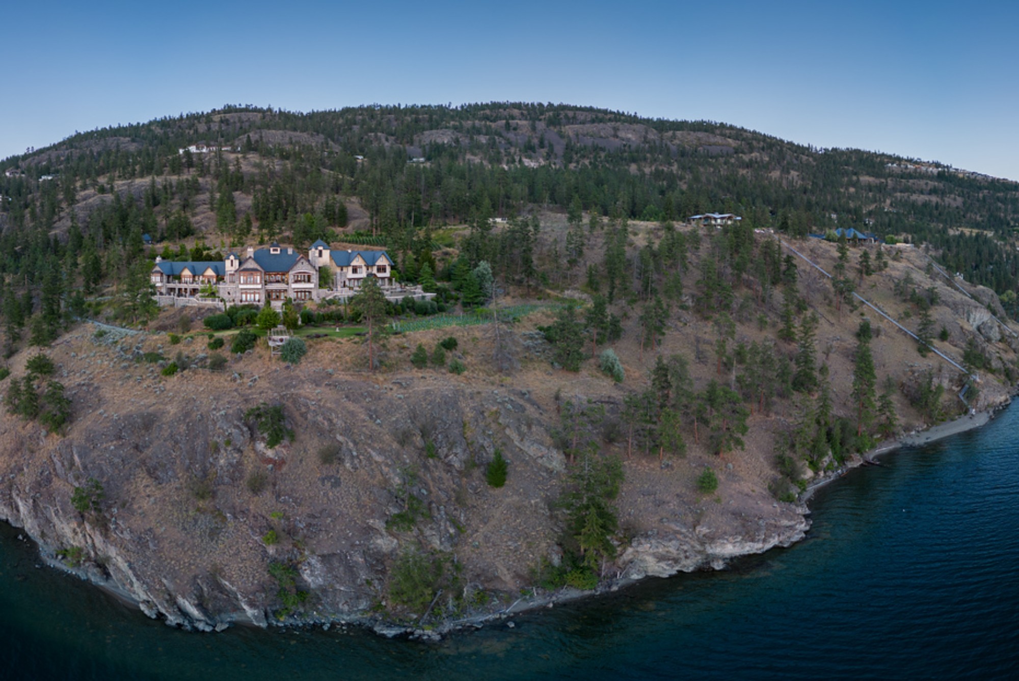 Aerial view of a luxurious waterfront estate surrounded by trees and rocky terrain, overlooking blue water.
