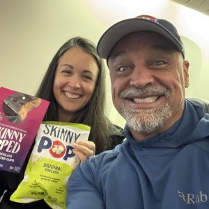 Two people smiling and holding snacks, including SkinnyPop original popcorn and chocolate peanut butter cups, on an airplane.