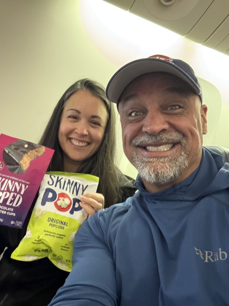 Two people smiling and holding snacks, including SkinnyPop original popcorn and chocolate peanut butter cups, on an airplane.