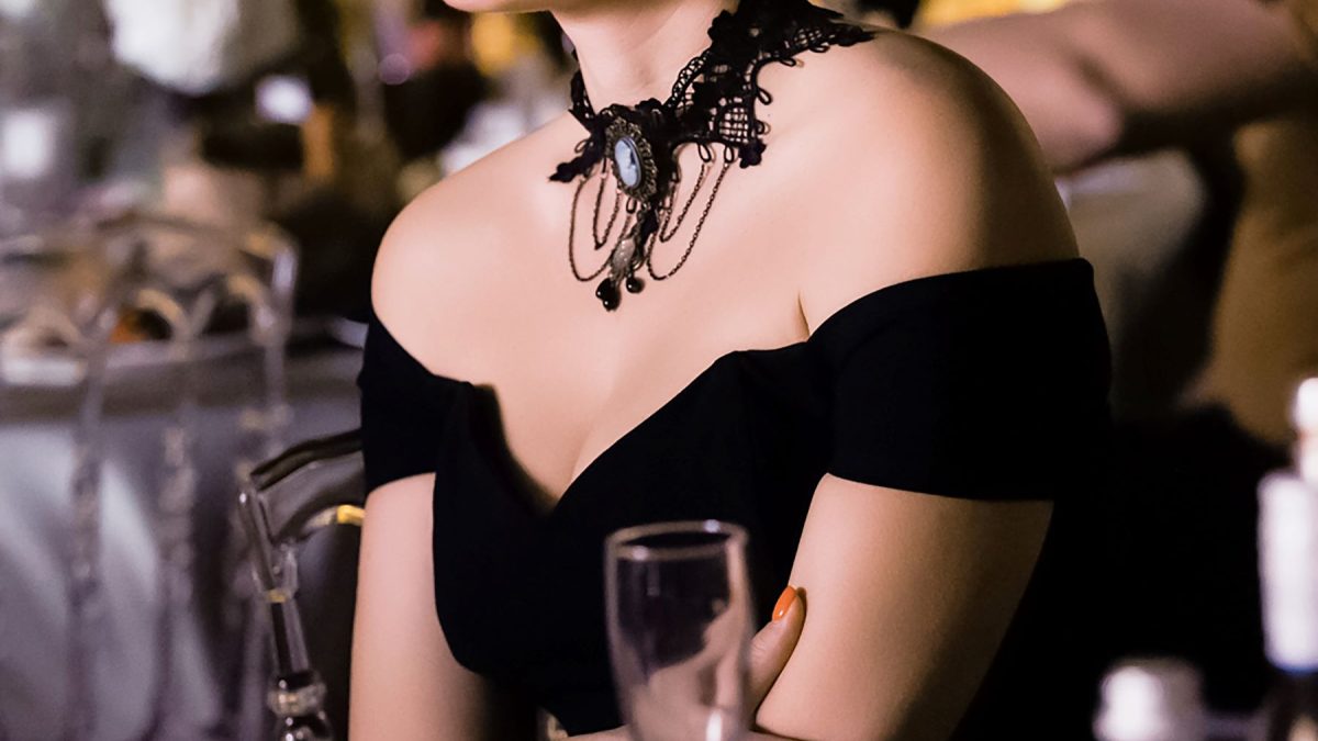 Elegant woman in a black off-shoulder dress with a decorative choker, seated at a luxurious venue with drinks nearby.