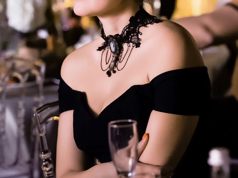 Elegant woman in a black off-shoulder dress with a decorative choker, seated at a luxurious venue with drinks nearby.